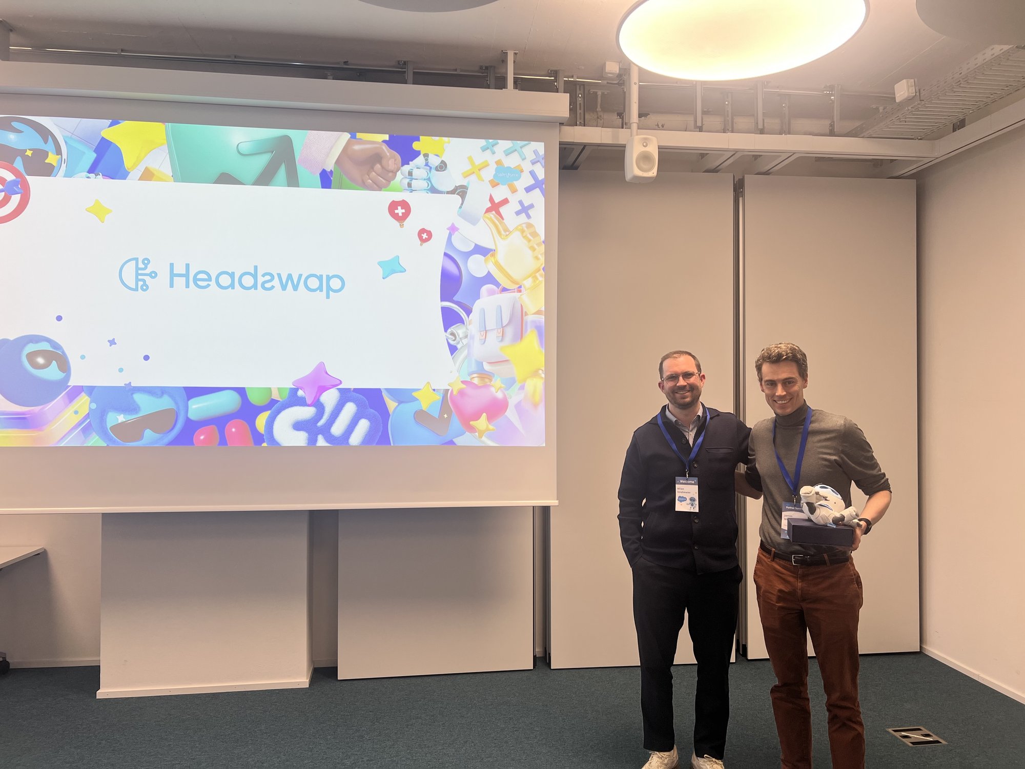 James and Milan at the Salesforce partner kickoff in Zurich, holding a Salesforce trophy in front of a Headswap-branded screen.